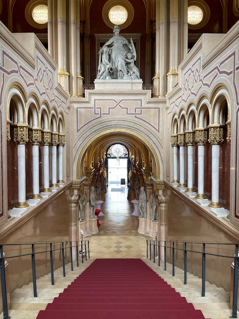 A grand stairway with a red carpet leading down to a polished marble floor, flanked by ornate white and gold columns supporting arches. Above, a large classical sculpture of a seated figure is positioned on a pedestal, with intricate decorative moldings and a circular window providing natural light. The building's interior features high ceilings, elegant architectural details, and a symmetrical layout, all exhibiting a clean and well-maintained appearance. This space appears to be part of a historic or civic building, with a polished, dust-free surface reflecting the ambient light. Carpet Cleaners Haringey might offer services to maintain such fine surfaces through professional cleaning and deep surface sanitisation.