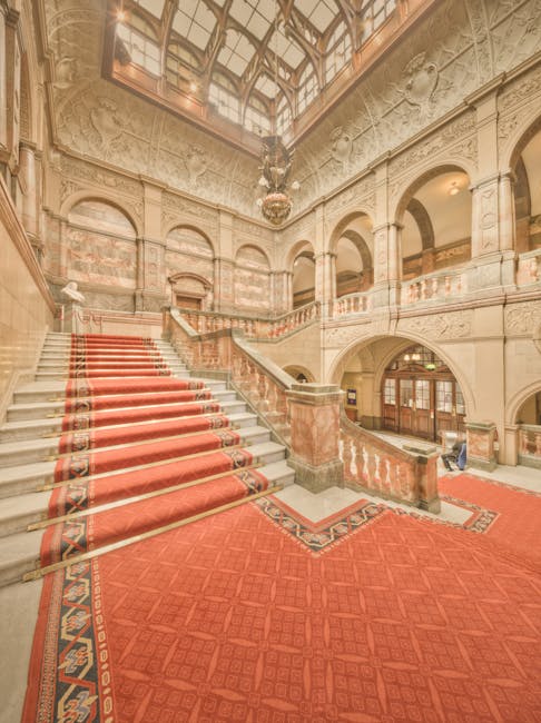 The image depicts the grand interior of Alexandra Palace featuring a large staircase with red carpet runners, flanked by ornate stone balustrades and intricate wall carvings. The polished marble surfaces and decorative mouldings showcase classic architectural detailing, illuminated by natural light filtering through a large glass-domed ceiling. The surrounding floors are covered with matching red carpet, and the spacious, high-ceilinged hall appears clean and well-maintained. Visible elements include the elegant staircase, stone columns, and decorative wall panels, contributing to the historic and opulent ambiance. This setting illustrates a space that benefits from routine deep cleaning and surface sanitisation, aligning with professional cleaning standards provided by Carpet Cleaners Haringey.