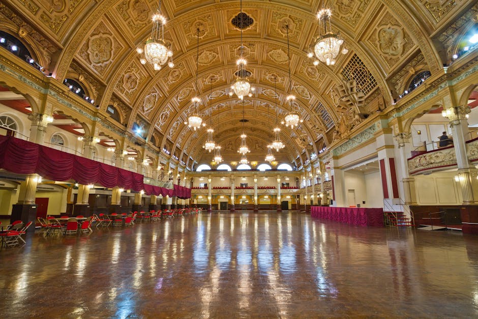 A grand stairway with a red carpet leading down to a polished marble floor, flanked by ornate white and gold columns supporting arches. Above, a large classical sculpture of a seated figure is positioned on a pedestal, with intricate decorative moldings and a circular window providing natural light. The building's interior features high ceilings, elegant architectural details, and a symmetrical layout, all exhibiting a clean and well-maintained appearance. This space appears to be part of a historic or civic building, with a polished, dust-free surface reflecting the ambient light. Carpet Cleaners Haringey might offer services to maintain such fine surfaces through professional cleaning and deep surface sanitisation.