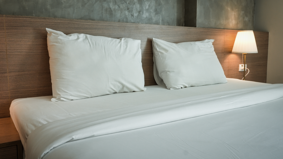 A modern bedroom featuring a neatly made bed with a white, textured mattress, a gray upholstered headboard, and minimalistic decor. The room has smooth white walls, a built-in beige wardrobe with a da
