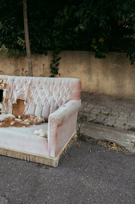 Exterior view of a pink upholstered sofa placed on a paved sidewalk next to a rough concrete wall and a narrow strip of outdoor paving. The sofa's fabric is heavily soiled and torn, with visible stuff