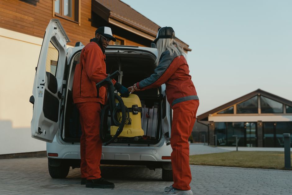 The open rear of a white commercial van parked on a paved driveway, showing various cleaning equipment and supplies inside. Visible surfaces include the black interior flooring and side panels of the 