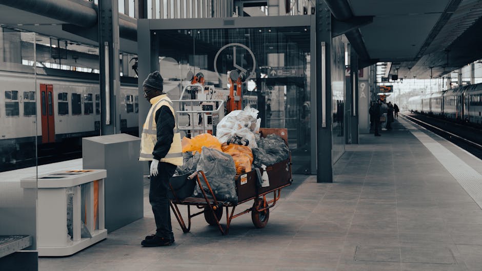A worker wearing a dark uniform, gloves, and a grey beanie hat, standing next to a heavily loaded wheeled trolley filled with trash bags and debris, on a modern train station platform with glass walls, metal structural elements, and a train parked on the adjacent tracks. The platform is clean and well-lit with natural light, and the worker appears to be engaged in janitorial or cleaning-related tasks. The scene is associated with Carpet Cleaners Haringey's comprehensive cleaning services, emphasizing surface sanitation and maintenance in public spaces.