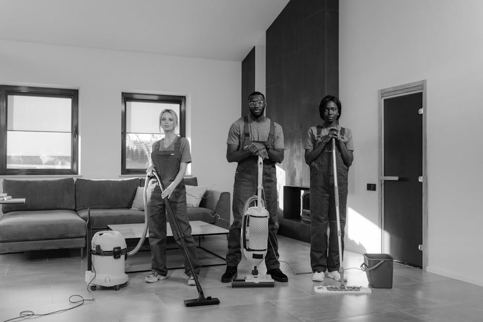 Three professional cleaners from Carpet Cleaners Haringey are performing surface cleaning and deep cleaning tasks in a modern residential living room. The room features a wooden floor being vacuumed by a woman on the left, a man in the center operating a steam or wet extraction carpet cleaning device, and a woman on the right holding a mop handle, with a bucket nearby. The room is illuminated with natural light coming through two large windows, which have minimal window treatments. The space includes a grey fabric sofa, a small coffee table, a corner cabinet, and a contemporary fireplace, all appearing well-maintained and clean. The walls are painted white, with one accent wall in a dark shade of paneling. Tools and cleaning equipment, such as a vacuum, mop, and cleaning sprays, are visible, indicating thorough sanitation and maintenance, aligning with the residential cleaning services offered by Carpet Cleaners Haringey to ensure hygienic, freshly cleaned surfaces.