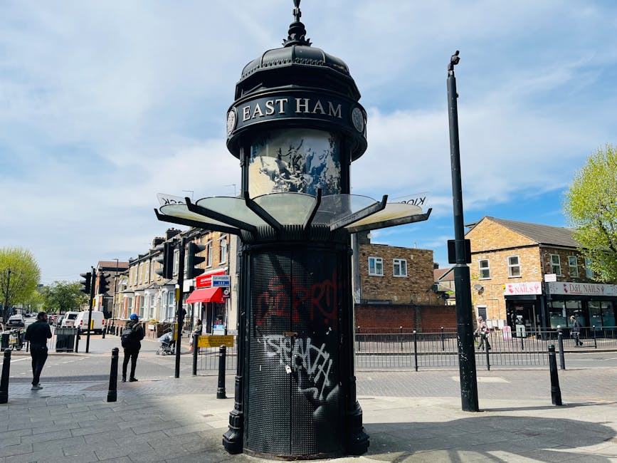 An outdoor public noticeboard and advertising kiosk on Tottenham High Street, Haringey, featuring a cylindrical metal structure with graffiti-style stickers and spray paint. The kiosk has a domed black top with the words 'East Ham' displayed around its circumference, and is surrounded by black metal fencing. In the background, there are residential and commercial buildings made of brick with shopfronts, including a nail salon, under partly cloudy blue skies. The area is busy with pedestrians walking along the paved sidewalk, and trees with fresh green leaves line the street, contributing to a vibrant urban scene captured during daytime.