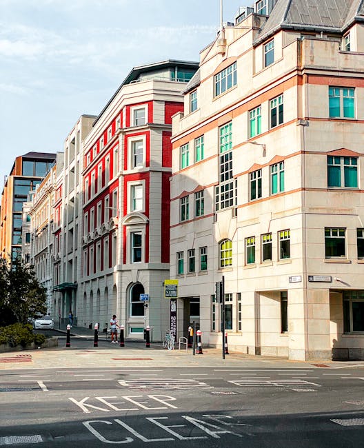 Photograph of a city street scene featuring modern multi-story office buildings with large windows, some framed in white and others in green, set against a clear blue sky. The buildings have a combination of brick and stone facades, with well-maintained exteriors. The street includes markings such as 'KEEP CLEAR' painted on the asphalt, with bollards, bicycle parking, and pedestrians visible. The overall scene emphasizes urban architecture and cleanliness, consistent with the professional cleaning and surface maintenance services provided by Carpet Cleaners Haringey, as showcased in their Tottenham high street office cleaning services page.
