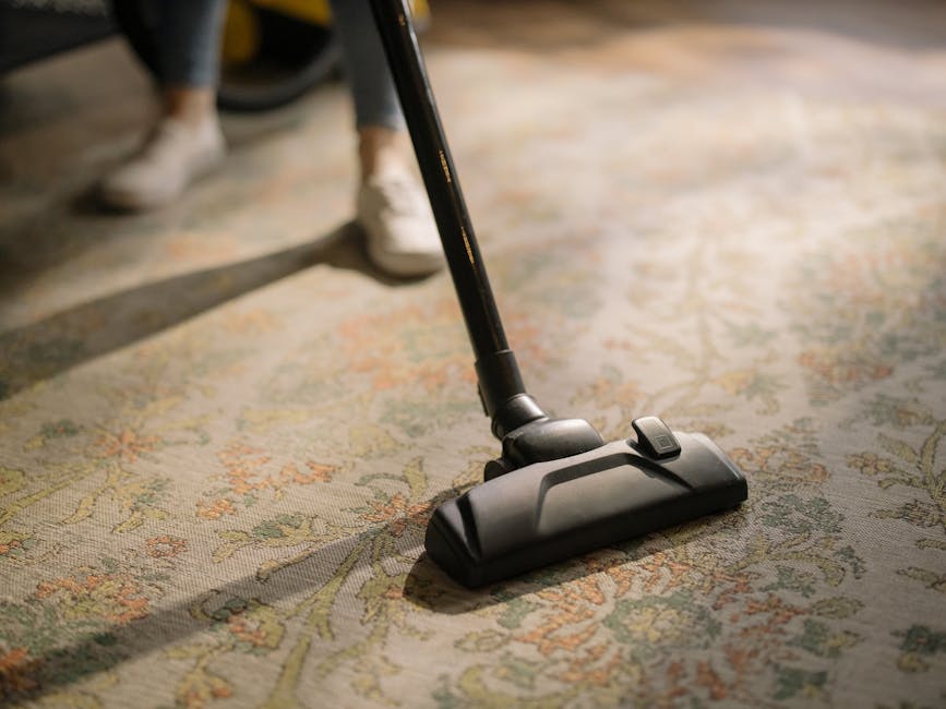 A young boy using a vacuum cleaner to clean a beige carpet in a living room with natural daylight coming through large windows, while an older man with gray hair and a beard is lying on a white sofa, 