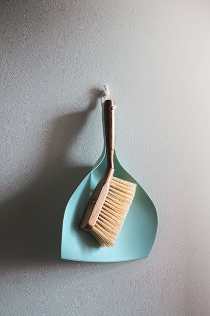 A light blue dustpan and a wooden-handled broom with beige bristles hanging on a white wall, illuminated by soft natural light. The cleaning tools are positioned with the broom's handle resting inside the dustpan, demonstrating a tidy and organized storage of cleaning equipment typically used in domestic or commercial surface cleaning. The background wall is smooth with a subtle texture, and the overall scene suggests readiness for deep cleaning tasks as part of a comprehensive end of tenancy cleaning service. This image reflects professional cleaning practices as offered by Carpet Cleaners Haringey, emphasizing hygiene and maintenance in residential or rental properties.