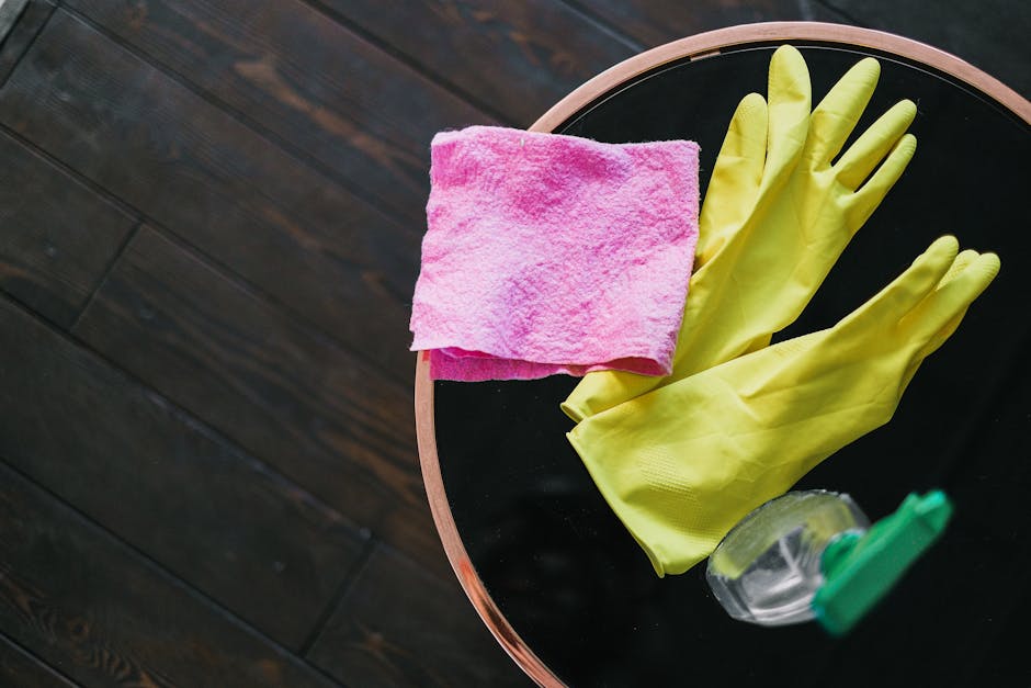 A round black tabletop with a pink microfiber cloth, a pair of yellow rubber gloves, and a small spray bottle of cleaning solution arranged neatly on its surface. The table is positioned next to dark wooden flooring, which is clean and well-maintained. The lighting in the room highlights the shiny surface of the tabletop and the vivid colors of the cleaning supplies. This setup emphasizes surface cleaning and sanitisation tools used in domestic cleaning offered by Carpet Cleaners Haringey, situated in the Wood Green end of tenancy cleaning guide N22 Haringey.